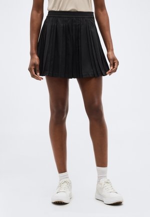 Άτομο που φοράει μαύρη πλισέ φούστα, λευκές κάλτσες και λευκά αθλητικά παπούτσια, στέκεται μπροστά σε ένα μονόχρωμο γκρι φόντο.