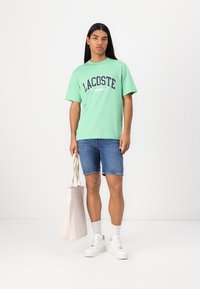 Hele roheline lühikeste varrukatega T-särk, millel on "LACOSTE" graafika, koos siniste teksashortsidega, valgete jooksujalatsitega ja beeži tooni kottiga.