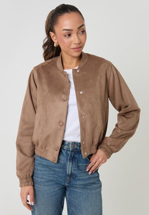 Femme portant une veste bomber en daim marron boutonnée sur une chemise blanche avec un jean bleu, posant avec une main sur la hanche et regardant vers la droite.