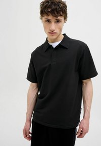 Polo noir en tissu texturé avec un col boutonné, des manches courtes et une étiquette logo sur le côté. Design simple avec une coupe décontractée.