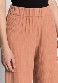 Pantalons larges plissés couleur pêche avec une taille élastique. Texture douce avec un détail en relief vertical. Présente une coupe décontractée.