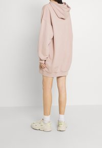 Rosa übergroßes Hoodie-Kleid mit Kapuze, langen Ärmeln, gerippten Bündchen und Saum. Kombiniert mit hellbeigen Sneakern und weißen Knöchelsocken.