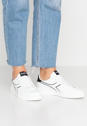 Hvide sneakers med sorte accenter, teksturerede sider og perforeringer. Parret med lyseblå cropped denim med tætsiddende kanter.
