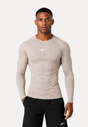 Atletisk mand iført en tætsiddende langærmet beige skjorte og sorte shorts, stående med hænderne i lommerne mod en ensfarvet baggrund.