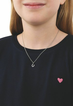 Joven que lleva un collar con un herradura plateada y una camiseta negra acanalada con un pequeño corazón rosa bordado en el pecho.