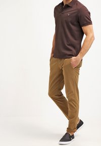 Muž v tmavě hnědém polo tričku, pískových chinos kalhotách s roll-up na kotnících a černých slip-on botách na jednobarevném bílém pozadí.