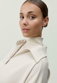 Moteris su atgal šukuota šukuosena, vilkinti aukštu kaklu smėlinę paltą, šiek tiek atsisukusi per petį prieš paprastą šviesią fono.