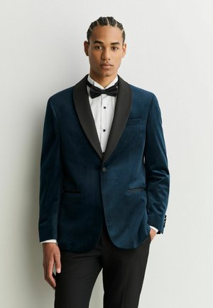 Young man with braided hair wearing a dark blue velvet tuxedo jacket, white shirt, black bow tie, and black pants standing against a white wall.
