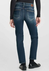 Jean en denim bleu foncé, coupe Slim, avec poches arrière, légères décolorations et coutures contrastées. Porté avec des chaussures et des chaussettes noires.