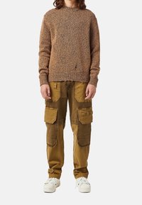 Maglione a maglia marrone con un motivo testurizzato, abbinato a pantaloni cargo beige con tasche e dettagli di pannelli a contrasto. Sneakers bianche.