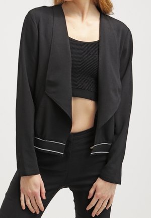Žena v černém saku s otevřeným předním zapínáním a bílým lemováním, který má přes texturovaný černý crop top a černé kalhoty s vysokým pasem.