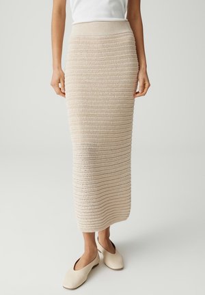 Donna che indossa una gonna midi beige a maglia, top bianco senza maniche e scarpe basse color crema, in piedi davanti a uno sfondo semplice.