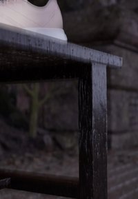 White sneaker resting on the edge of a dark wooden bench with a blurred stone wall and tree in the background.
