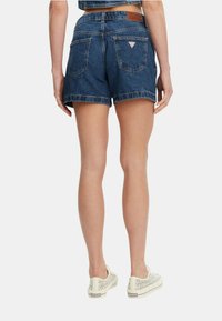 Shorts in denim blu scuro con orli sfrangiati, dotati di tasche posteriori e un'etichetta in pelle del marchio. Abbinati a sneakers chiare.