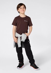 Braunes Baumwoll-T-Shirt mit Logo, graues Sweatshirt um die Taille gebunden, schwarze Jogginghose und schwarze Sportschuhe mit roten und weißen Akzenten.