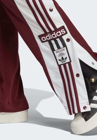 Pantalones deportivos burdeos con paneles blancos que presentan tres rayas, botones de presión y un parche con el logotipo de adidas; combinados con zapatos negros.