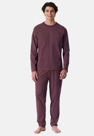 Man staat blootsvoets, draagt een bijpassende top met lange mouwen en borstzak en een broek in donker mauvekleur, lacht naar de camera.