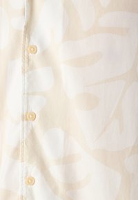 Primo piano di un tessuto beige e bianco a fantasia con tre bottoni beige chiaro cuciti verticalmente lungo una cucitura.