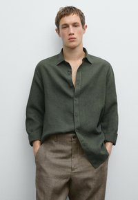 Dark green linen shirt with a button-up front, chest pocket, and rolled cuffs, paired with brown textured trousers.