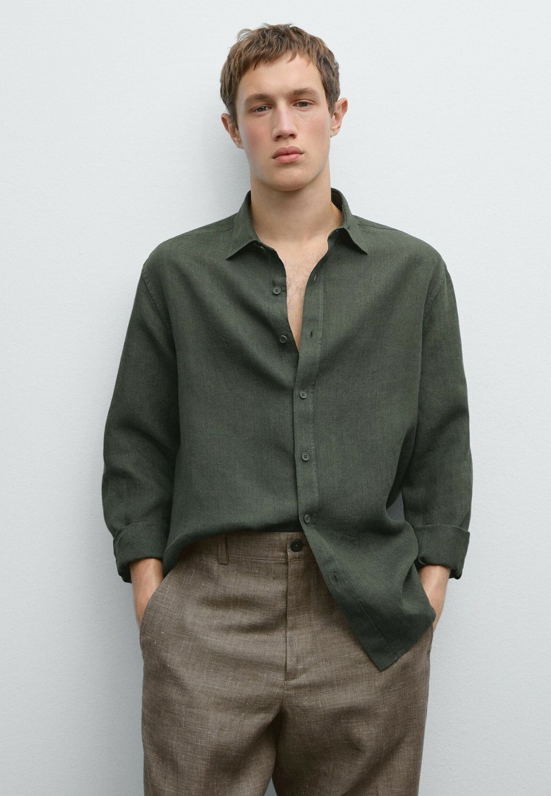 Dark green linen shirt with a button-up front, chest pocket, and rolled cuffs, paired with brown textured trousers.