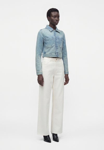 Γυναίκα όρθια, φοράει ένα ανοιχτό μπλε cropped denim μπουφάν, λευκό ψηλόμεσο παντελόνι με φαρδιά πόδια και μαύρες μυτερές μπότες.