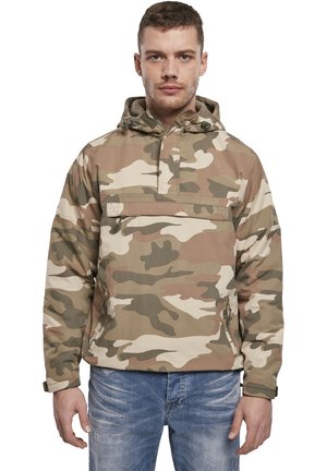 Chaqueta de camuflaje con capucha, hecha de tejido ligero. Presenta un bolsillo frontal y puños elásticos. La paleta de colores incluye tonos beige y verde.