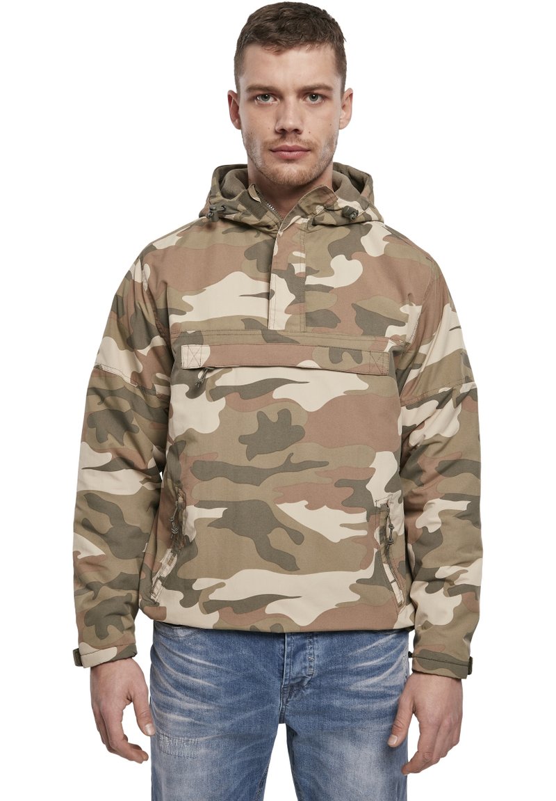 Veste camouflage avec capuche, fabriquée en tissu léger. Présente une poche avant et des poignets élastiques. La palette de couleurs comprend des tons beige et vert.