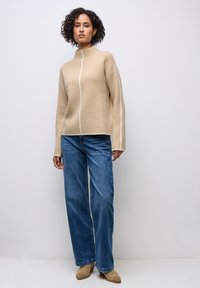Beige Wollmischpullover mit hohem Kragen und Mittelnaht, kombiniert mit weit geschnittenen blauen Denim-Jeans und beigen Knöchelstiefeln.