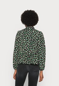 Blusa preta com um padrão de bolinhas verdes e brancas, mangas longas, cintura ajustada e um design de peplum. Tecido suave com uma textura leve.