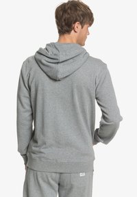 Sudadera gris de tela suave con capucha ajustable mediante cordón, puños y dobladillo acanalados. Presenta una pequeña etiqueta de logo cerca de la cintura, vista trasera mostrada.