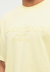 Vyras dėvi šviesiai geltoną marškinėlį su tekstūra, kuriame parašyta "TOMMY JEANS nuo 1985 metų Niujorko mieste" ant krūtinės.