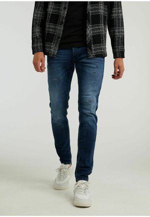 Mężczyzna ubrany w ciemnoniebieskie, dopasowane, przetarte dżinsy, białe trampki, czarną koszulę oraz czarno-białą kurtkę w kratę, stojący na jednolitym tle.