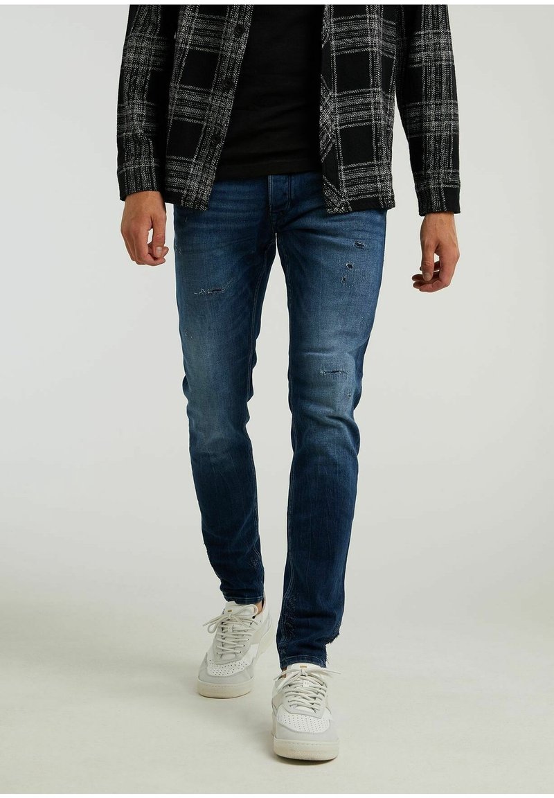 Homme portant un jean slim déchiré bleu foncé, des baskets blanches, une chemise noire et une veste à carreaux noir et blanc, debout sur un fond uni.