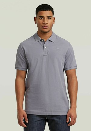 Jonge man die een lichtgrijs poloshirt met korte mouwen en donkerblauwe spijkerbroek draagt, staand tegen een effen lichte achtergrond.