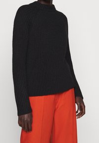Schwarzer gerippter Strickpullover mit hohem Kragen und langen Ärmeln, kombiniert mit leuchtend orangen weit geschnittenen Hosen. Strukturiertes Material und lockere Passform.