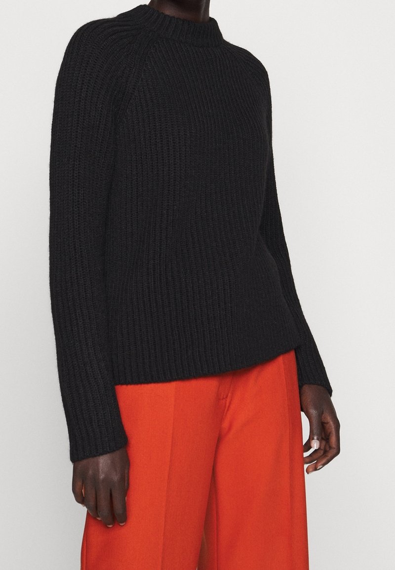 Schwarzer gerippter Strickpullover mit hohem Kragen und langen Ärmeln, kombiniert mit leuchtend orangen weit geschnittenen Hosen. Strukturiertes Material und lockere Passform.
