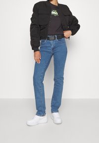 Giacca bomber nera con tasche frontali, t-shirt scura con grafica, jeans blu a gamba dritta, sneakers bianche e cintura nera visibile.