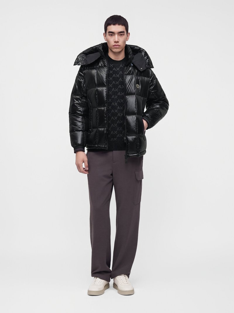 Jeune homme portant une doudoune noire brillante, un pull noir à motif, un pantalon cargo gris foncé et des baskets blanches, debout devant un fond blanc.