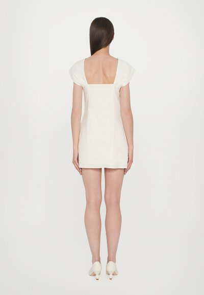 White linen dress with short puff sleeves, square back, and fitted silhouette, paired with cream-colored low-heeled shoes.