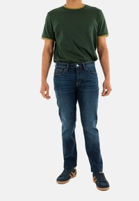 T-shirt verde con rifiniture gialle, jeans in denim blu e sneaker scuri. Jeans slim-fit con tasche, lieve scolorimento sul denim.