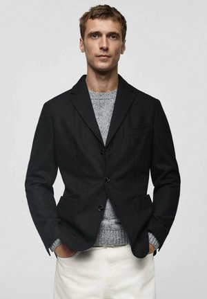 Black blazer with two front pockets, single-breasted design, paired with a light gray sweater and cream-colored trousers. Flat textures and clean lines.