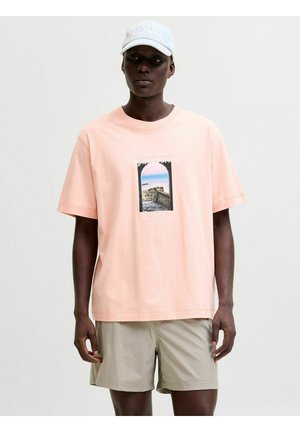 Personne portant un t-shirt graphique rose clair avec un imprimé arche en bord de mer, un short beige et une casquette de baseball blanche sur fond uni.