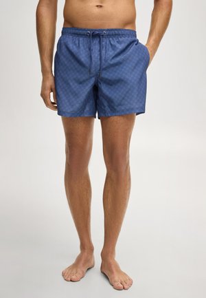 Homme portant un short de bain bleu à motifs avec un cordon de serrage, debout pieds nus devant un fond uni.