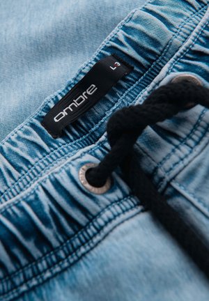 Nahaufnahme des Bunds einer hellblauen Jeanshose mit schwarzem Kordelzug und schwarzem Größenetikett mit der Aufschrift "ombre L".