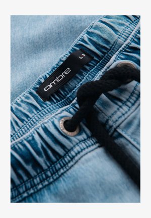 Close-up van de tailleband van lichtblauwe spijkerbroek met zwarte trekkoord en zwarte maatlabel met de tekst "ombre L".