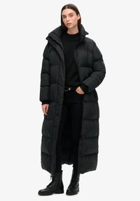 Lång svart puffjacka med hög krage, vadderad design, tryckknappar och en avslappnad passform, bärs över svarta kläder och kängor.