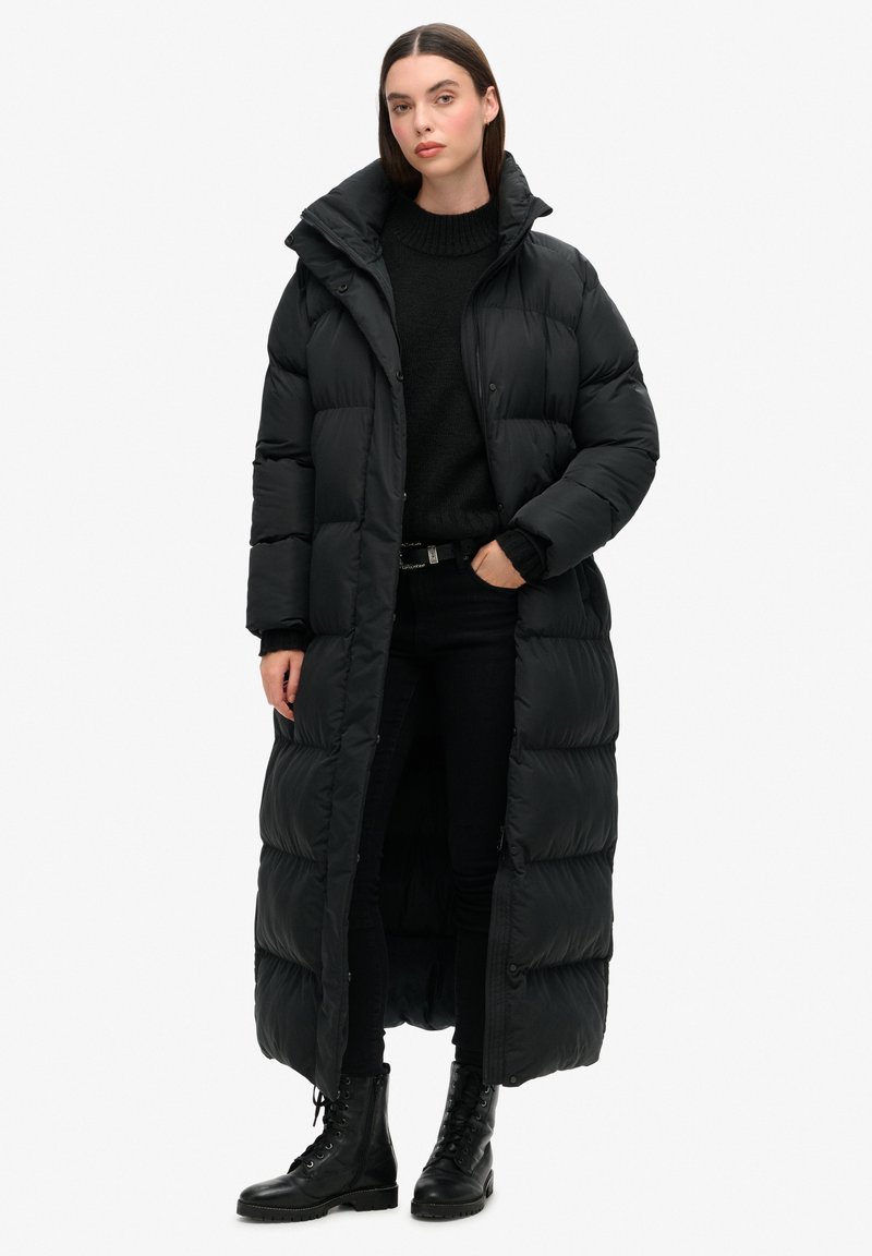 Abrigo puffer largo negro con un alto cuello, diseño acolchado, botones a presión y un corte holgado, llevado sobre ropa negra y botas de combate.