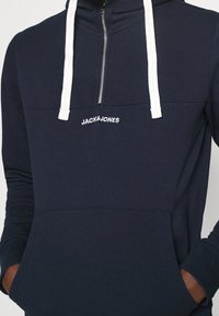 Námořnická fleecová mikina s bílými šňůrkami, předním polozipem a klokaní kapsou. Na hrudi je vyšitý nápis "JACK & JONES".