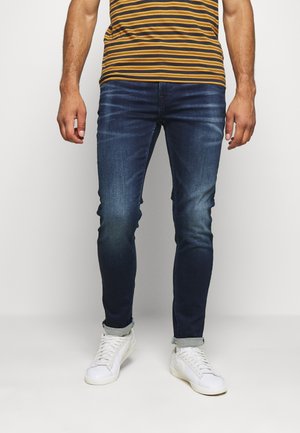 Man die donkerblauwe slim-fit jeans, een geelbruine gestreepte shirt en witte sneakers draagt, staand tegen een effen achtergrond.