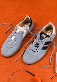 Baskets en daim bleu clair avec des rayures noires et une étiquette "CAVAL", lacets défaits, posées sur une surface texturée orange à côté d'un casque orange.